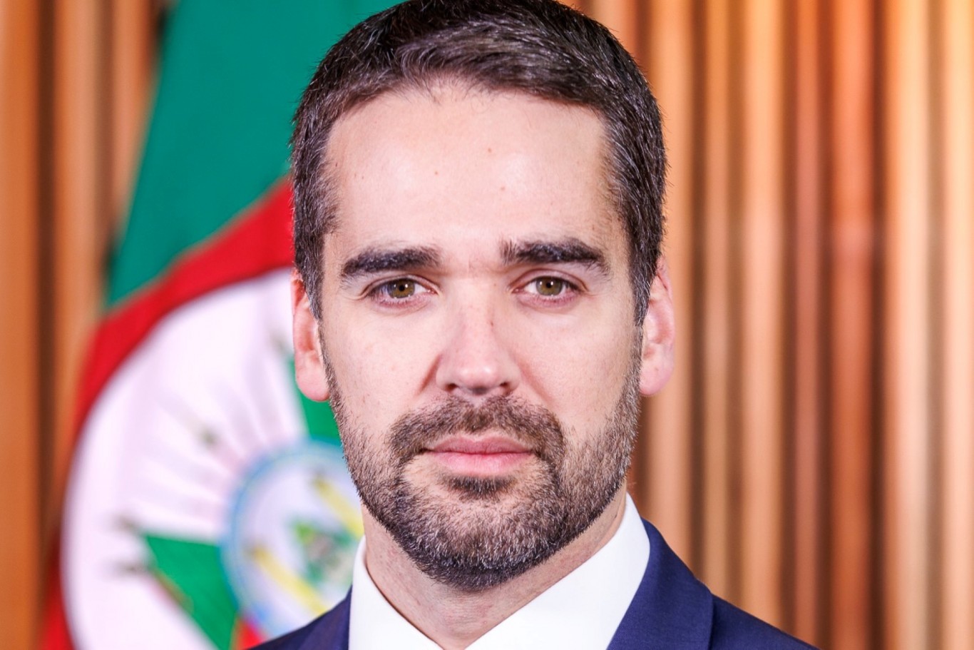 Retrato do governador Eduardo Leite, com uma bandeira do RS ao fundo. É um homem jovem cabelo curta castanho, olhos também castanhos, de barba aparada.